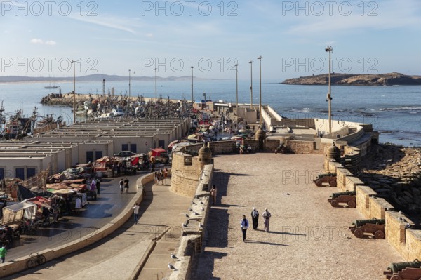 Harbour with lively market activity next to historic walls under clear skies