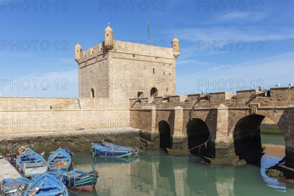 Old stone bridge with small colorful boats in a quiet harbor and a historic fortress in the background