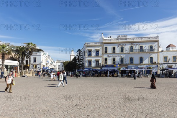 People walk on a wide square surrounded by historic buildings in clear skies