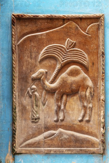 Wood carving of a camel depicting a person in traditional clothing against a hill backdrop, traditional door design