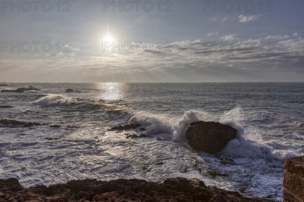 Waves crash against rocks in the ocean at sunset under a cloudy sky