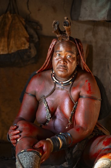 Elderly Himba woman rubbing herself with red paste for skin care, in a hut, traditional Himba village, Kaokoveld, Kunene, Namibia