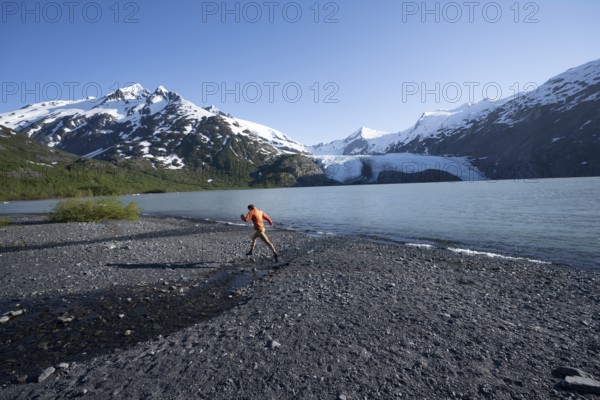Young man jumping on a pebble beach on a glacial lake, Snowy Mountains and Portage Glacier on Portage Lake glacial lake, Chugach National Forest, Alaska, USA