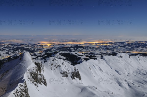 Night shot, long exposure of the 2506 meter high Säntis into the wintery, snow-covered, fog-covered foothills of the Alps illuminated by the villages, during a clear full moon night, Canton of Appenzell, Switzerland