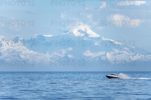 View over Cook Inlet to white mountain peaks of Mount Iliamna, motor boat rides on the ocean, mountains of the Aleutian Range, Anchor Point, Anchor River State Recreation Area, Alaska, USA