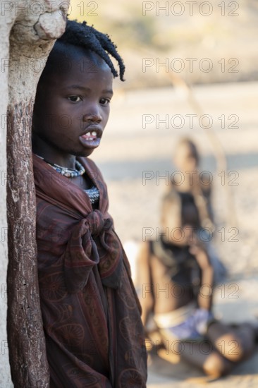 Himba girl leaning at a traditional clay hut, in the morning, traditional Himba village, Kaokoveld, Kunene, Namibia