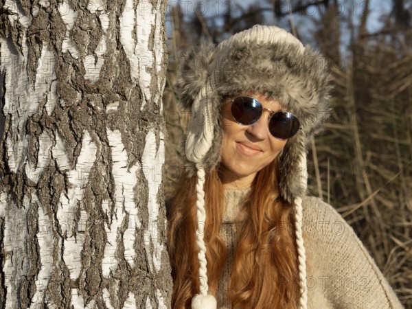 Slender pretty woman with fur hat and sweater, red hair and warm winter colors between birch forest and reeds