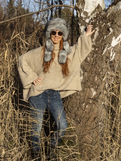 Slim woman with fur hat and sunglasses, red hair and warm winter colors between birch forest and reeds