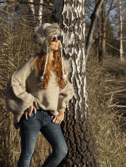 Slender pretty woman with fur hat and pompoms, red hair and warm winter colors between birch forest and reeds