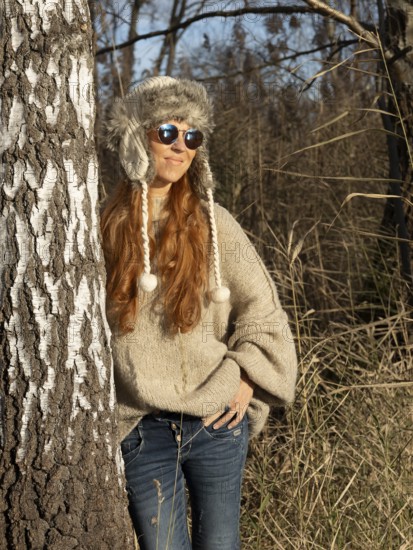 Slender pretty woman with fur hat and sweater, red hair and warm winter colors between birch forest and reeds