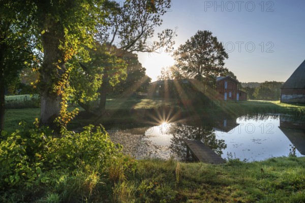 Manor with historic stables and pond, from 1923, in backlight with sun star, Gut Othenstorf, Mecklenburg-Western Pomerania, Germany