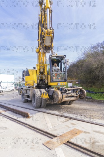 A yellow excavator on a construction site with tracks in the foreground. The sky is clear and the surrounding area is calm, track construction of the Hermann Hesse Railway, Calw, Germany