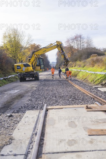 Construction workers lay tracks on a construction site while a yellow excavator assists. Autumn trees in the background, Hermann Hesse Railway track construction, Calw, Germany