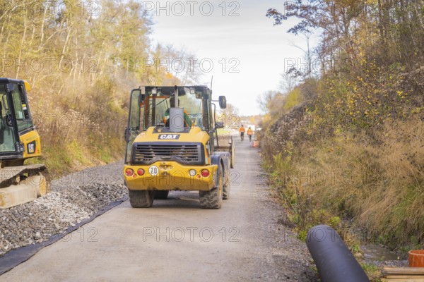 A yellow excavator is standing on a construction site, surrounded by autumn trees. Workers can be seen in the distance, track construction of the Hermann Hesse Railway, Calw, Germany