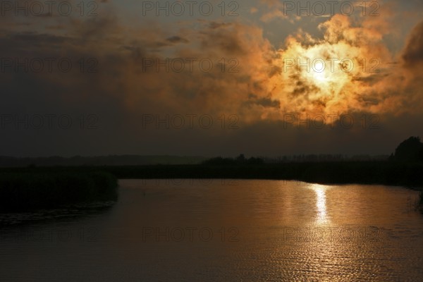 Dramatic sunset behind clouds over a quiet river with golden reflection, Peenetal nature park Park, Mecklenburg-Western Pomerania, Germany
