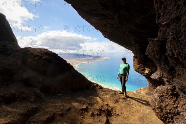 Tourist in the Cueva de las Cabras cave, young woman enjoying the view from the Risco de Famara cliff to Famara beach, Playa de Famara with La Calaeta, Lanzarote, Canary Islands, Spain