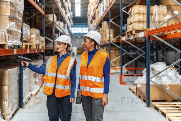 Two women wearing hard hats and safety vests are analyzing inventory by scanning product barcodes inside a modern logistics distribution warehouse with tall shelving racks