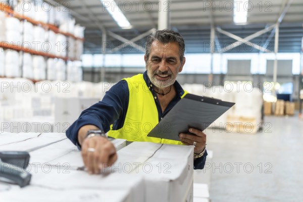 Logistics manager smiling while using a barcode scanner and clipboard checking shipment boxes in a large modern distribution warehouse, ensuring efficient supply chain operations