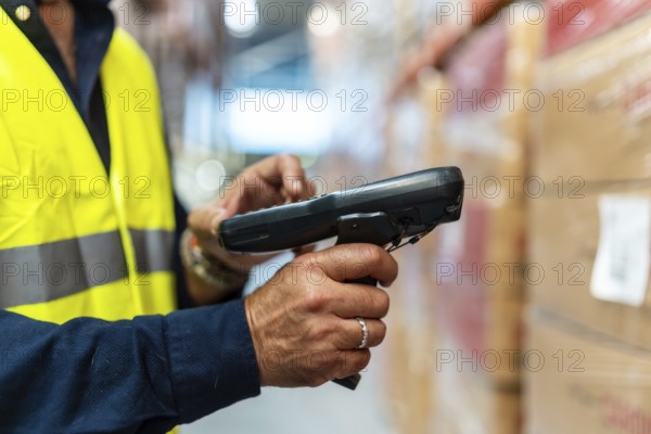 Warehouse worker in high visibility vest and blue uniform scanning barcodes on boxes with handheld scanner, updating inventory and tracking stock in a busy distribution center