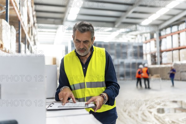 Experienced manager wearing a high visibility safety vest confirming details on a clipboard while standing in a busy logistics warehouse, overseeing stock and supply chain operations