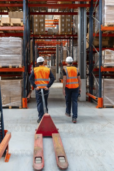 Warehouse workers in safety vests and hard hats navigating through industrial shelving with a pallet jack, managing stock and coordinating distribution within a large logistics center