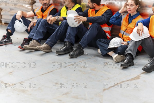 Group of diverse warehouse workers sitting on the floor taking a break, feeling exhausted after a long shift, wearing safety workwear in a logistics warehouse