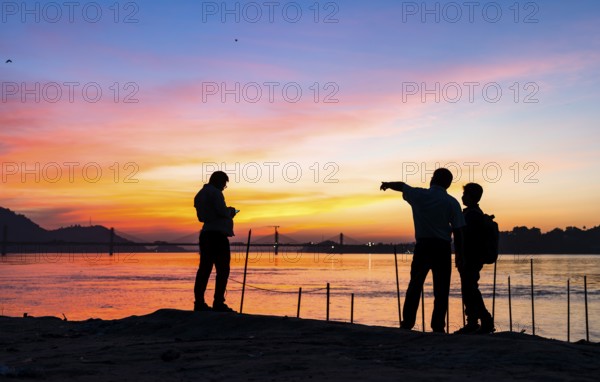 People enjoy the vibrant sunset by the Brahmaputra riverside, capturing photos and moments against the scenic backdrop of a bridge and glowing sky, on November 4, 2025 in Guwahati, India