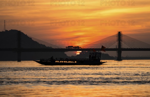 A boat sails on the river Brahmaputra during sunset, with the silhouette of a cable-stayed bridge and hills in the background, on November 4, 2025 in Guwahati, India