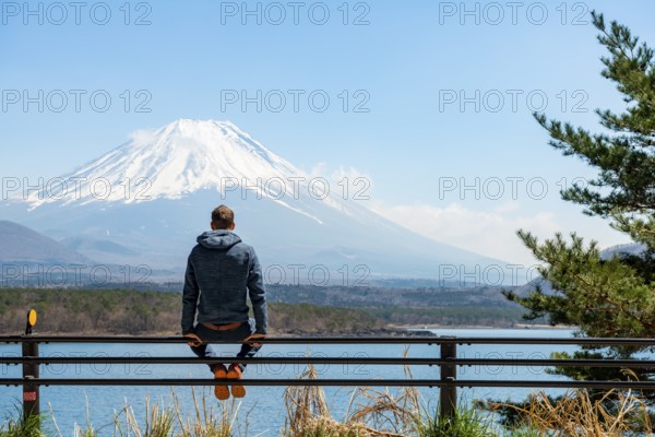 Young man sitting on railings next to a road and looking across the lake to Mt Fuji volcano, Motosu Lake, Yamanashi Prefecture, Japan