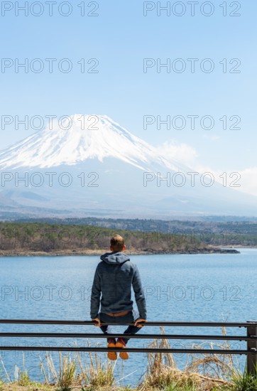 Young man sitting on railings next to a road and looking across the lake to Mt Fuji volcano, Motosu Lake, Yamanashi Prefecture, Japan