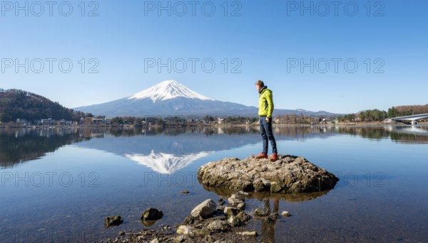Young man standing on a rock in water, volcano Mt. Fuji is reflected in Lake Kawaguchi, Yamanashi Prefecture, Japan