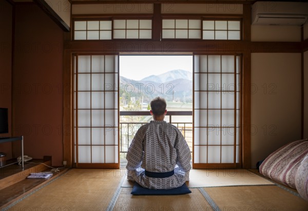 Young man wearing kimono sitting in traditional Japanese living room with tatami mats and shoji sliding doors, from behind, Yamanouchi, Nagano, Japan