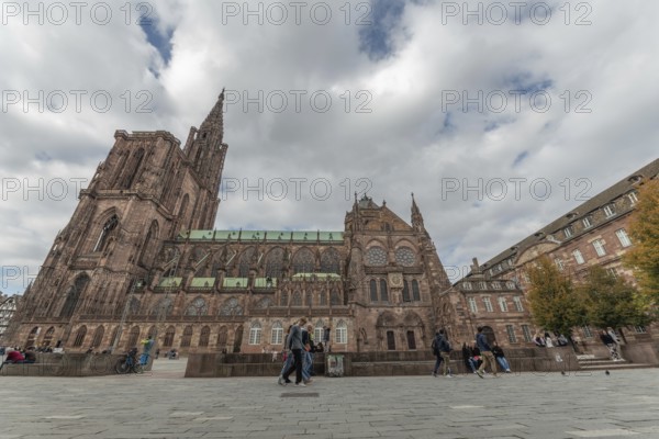 People walk in front of the majestic Strasbourg Cathedral, which is surrounded by historic buildings, on gray autumn Monday. The architecture attracts a lot of tourists. Bas Rhin, Alsace, France