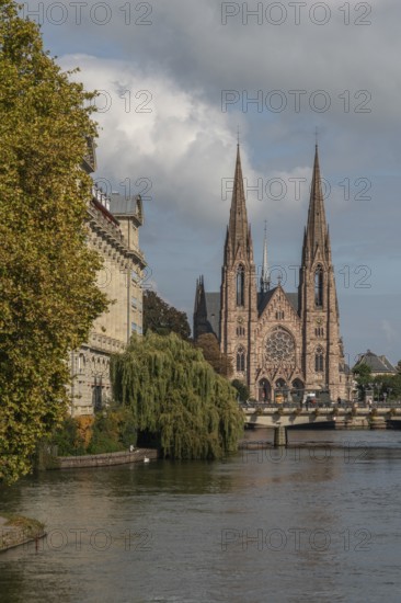 The two-towered church stands majestically on the banks of the river. Trees with golden leaves surround historic buildings under cloudless ski slopes. Bas Rhin, Alsace, France