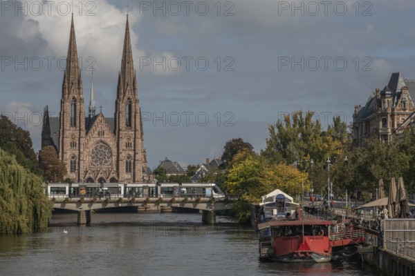 The two-towered church stands majestically on the banks of the river. Trees with golden leaves surround historic buildings under cloudless ski slopes. Bas Rhin, Alsace, France