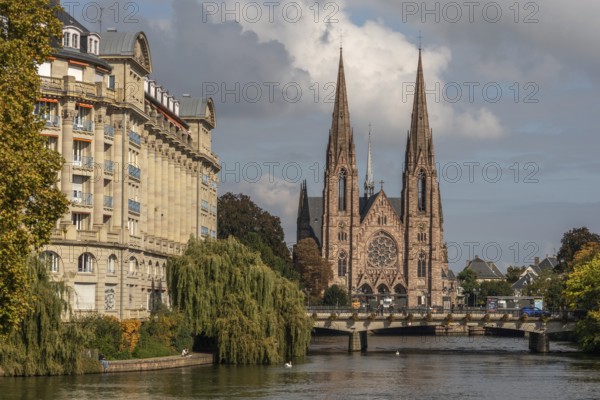 The two-towered church stands majestically on the banks of the river. Trees with golden leaves surround historic buildings under cloudless ski slopes. Bas rhin, Alsace, Grand Est, France