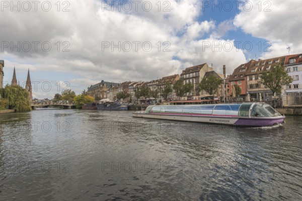 A pleasure boat glides peacefully on the Strasbourg River. Colourful buildings and trees line the banks and create a relaxing atmosphere. Bas Rhin, Alsace, France