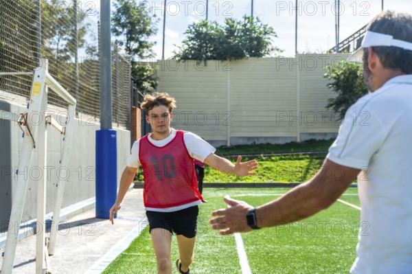 Youth male soccer player in a red bib running towards a coach with outstretched arms, receiving guidance during outdoor sports training on a green field