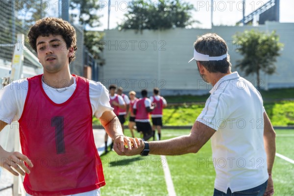 Coach instructing a soccer player on the field during training, teammates jogging in the background as the team practices teamwork, strategy and athletic development