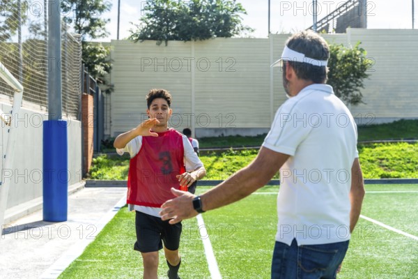 Youth male soccer player in a red bib running towards a male coach with an outstretched hand, receiving instruction during a training session on a green artificial turf field