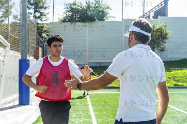 Soccer coach high fiving young male player in red bib on green artificial turf, offering encouragement and guidance during outdoor training practice, teamwork and mentorship