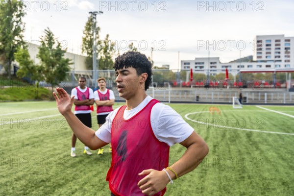 Soccer coach providing instructions to a group of young men in red bibs during a training session on a green artificial turf field from a close up perspective