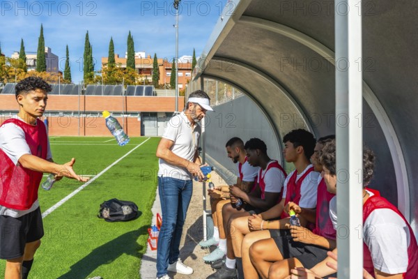 Soccer coach distributing water bottles to young male players resting in the dugout during a training session or match, emphasizing hydration and teamwork