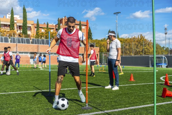 Soccer player is training on a professional artificial turf field, dribbling a ball through obstacles, with a coach observing team members during a group practice session