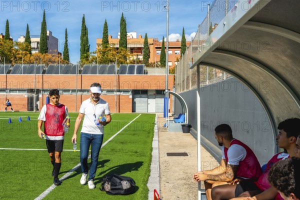 Soccer coach and young player walking on a green artificial turf field after training, carrying water bottles, with other team members resting in the dugout