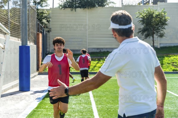 Soccer coach extending hand for a high five with a young male player wearing a red bib during a team training session on a green artificial turf field, with other players in background