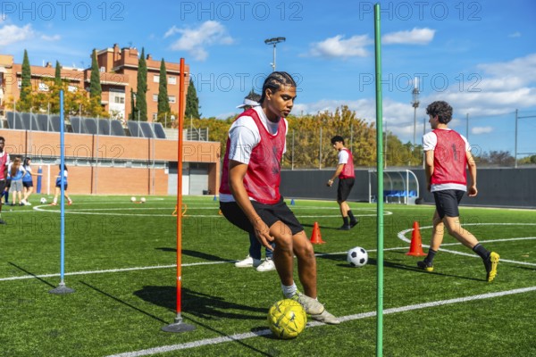 Young man in a red vest training soccer dribbling techniques on a green artificial turf field, navigating around cones and poles during an outdoor practice session with other players learning