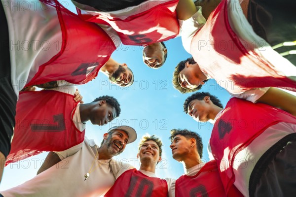 Coach and young diverse male soccer players forming a huddle, discussing strategy and fostering collaboration, looking down at them from below against a bright blue sky
