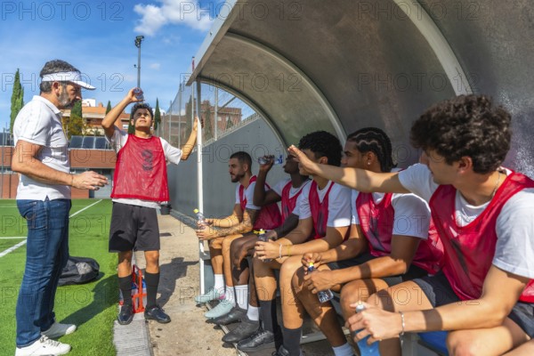 Soccer coach briefing male players on the sideline bench during a break, discussing strategy and hydration while the team rests and prepares to return to the match