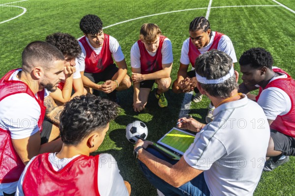 Soccer coach providing strategic instructions to his diverse team of young male players, gathered in a huddle on a green artificial turf field with a soccer ball nearby